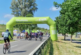 Ein grüner Startbogen der Veranstaltung „Rheinradeln“ steht an einer Radstrecke entlang des Rheins zwischen Worms und Oppenheim. Die Aktion lädt Radfahrende ein, die Region entlang des Flusses autofrei zu entdecken. © CC0, Katja Zentel Grüner aufblasbarer Startbogen mit der Aufschrift „Gemeinsam Zukunft anpacken – EWR Energie auf neuen Wegen“ über einer Radstrecke beim Rheinradeln zwischen Worms und Oppenheim. Im Hintergrund stehen Bäume unter blauem Himmel.