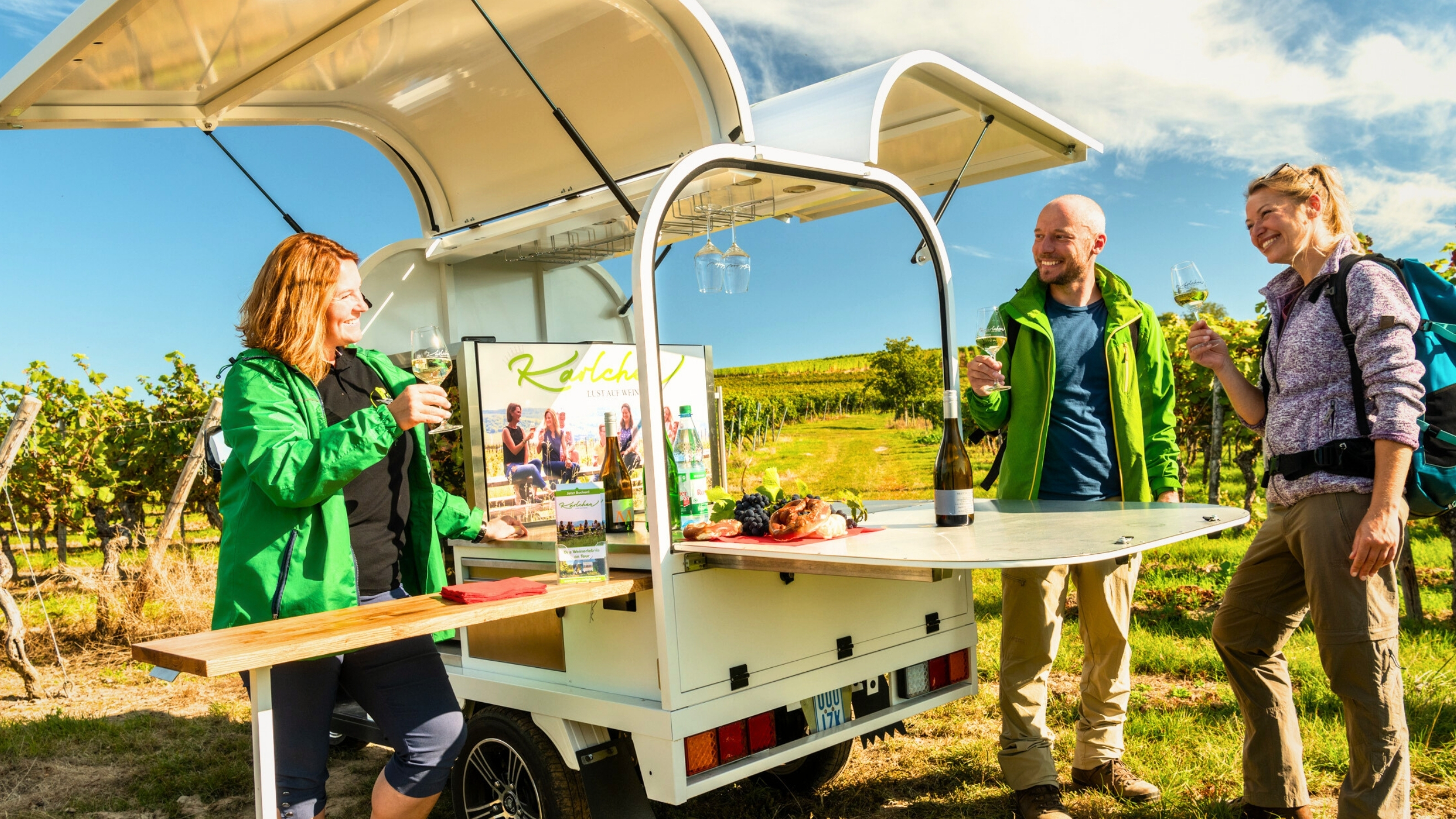 Drei Menschen stehen an einem kleinen mobilen Weinstand auf Rädern zwischen Weinreben. Eine Frau hinter dem Stand schenkt Wein aus und spricht mit zwei Wandernden, die jeweils ein Glas Weißwein in der Hand halten und Rucksäcke tragen. Auf dem Tresen stehen eine Weinflasche, Wasser, Trauben und Brot. Die Szene spielt an einem sonnigen Tag inmitten der Weinberge.