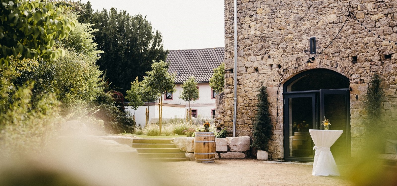 Rustikaler Hof mit Sandsteinmauer, Weinfass, Stehtisch und grüner Gartenumgebung – ideal für Veranstaltungen.