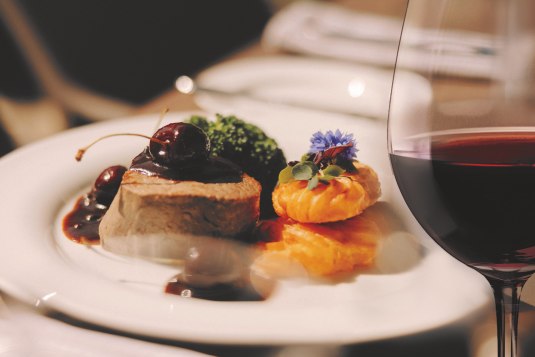 Feines Gourmetgericht mit Fleisch, Soße, Gemüse und Kartoffelrosetten – serviert mit einem Glas Rotwein.