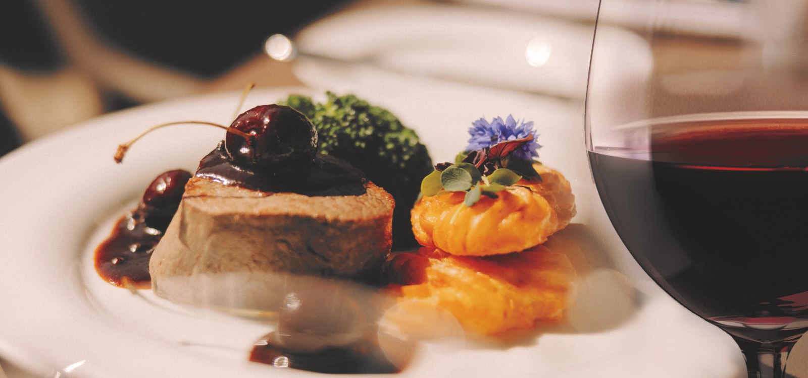 Feines Gourmetgericht mit Fleisch, Soße, Gemüse und Kartoffelrosetten – serviert mit einem Glas Rotwein.