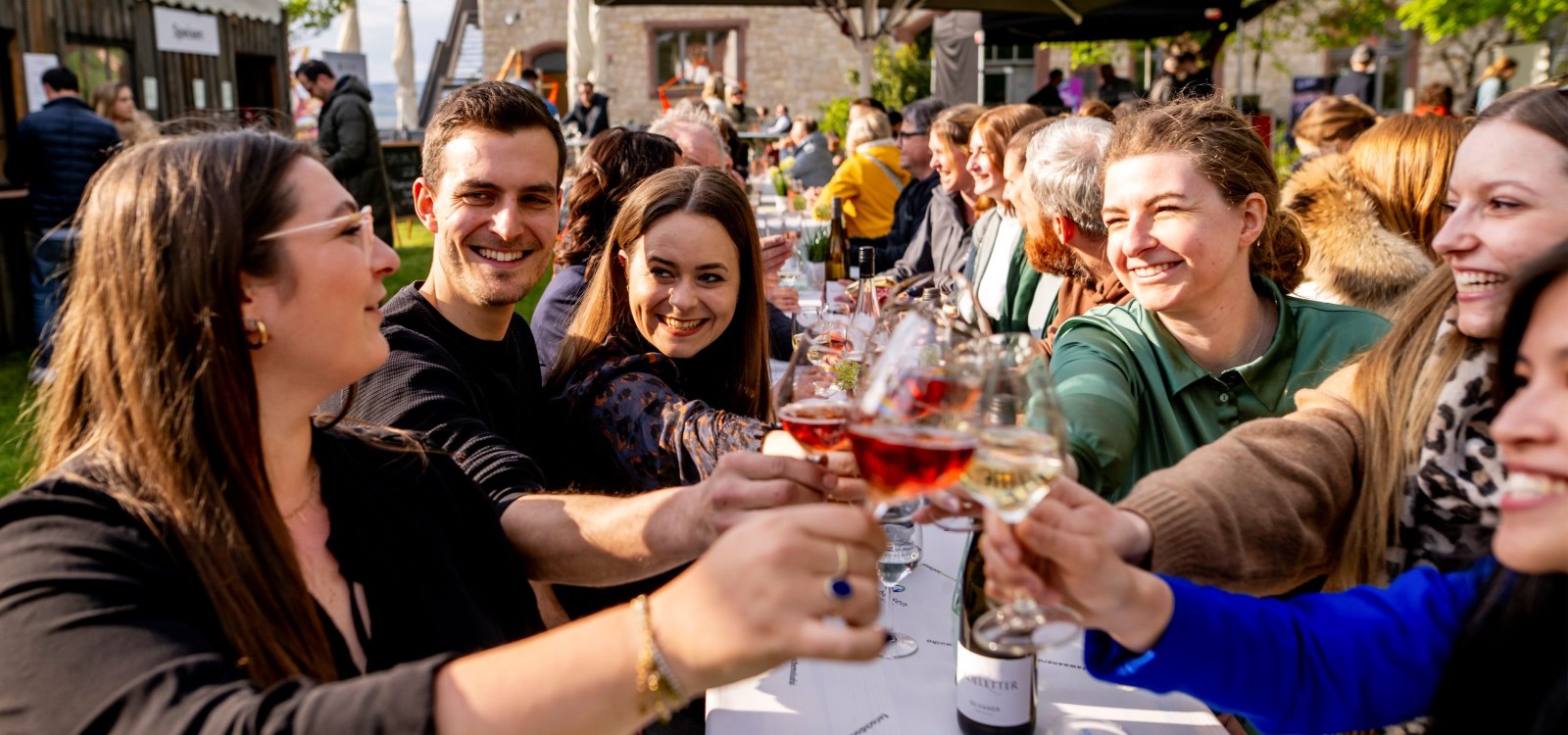 Mehrere Personen sitzen an einer langen Tafel im Freien und sto&szlig;en mit Weingl&auml;sern an. Im Hintergrund sind ein steinernes Geb&auml;ude, Sonnenschirme und weitere G&auml;ste beim Fr&uuml;hlingsfest in Ingelheim zu sehen.