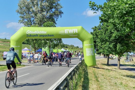 Groene opblaasbare startboog met de tekst &bdquo;Gemeinsam Zukunft anpacken &ndash; EWR Energie auf neuen Wegen&ldquo; boven een fietsroute tijdens Rheinradeln tussen Worms en Oppenheim. Op de achtergrond staan bomen onder een blauwe lucht.