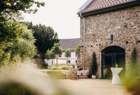 Rustikaler Hof mit Sandsteinmauer, Weinfass, Stehtisch und grüner Gartenumgebung – ideal für Veranstaltungen.