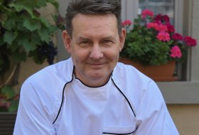 Porträt eines Kochs Alex Greiner mit Glas Rotwein im Hofambiente, umgeben von Weinreben und Blumen.